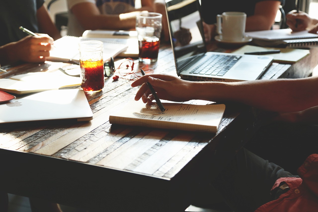 A table with a laptop, notepad, and hand holding a pencil to take notes.